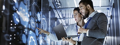 IT-Spezialist mit Laptop im Gespräch mit einem Servertechniker in einem Serverraum in einem Rechenzentrum mit Wolken- und Schildsymbolen auf den Serverschränken