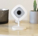 A white security camera sitting between a candle cup and a potted cactus.